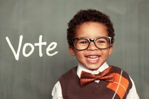 image: young African-American child with thick rimmed glasses, the word VOTE in background